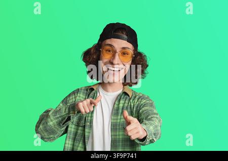 Lächelnder junger Mann mit lockigem Haar und Sonnenbrille zeigt auf die Kamera, positive Stimmung, Jugendkultur Stockfoto