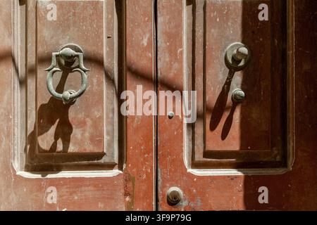 Nicht passender metallener Türklopfer und Griff werfen lange Schatten auf eine verwitterte rötlich-braune Holztür, Valletta, Malta Stockfoto