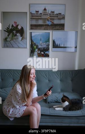 Junge Frau benutzt Smartphone, während sie zu Hause auf dem Sofa mit ihrer Katze sitzt, alltägliches digitales Lebensstilkonzept. Stockfoto