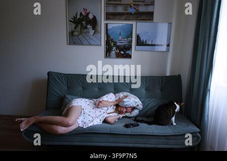Junge Frau liegt auf dem Sofa und bedeckt ihre Ohren mit Kissen, während die Katze daneben sitzt, Stress- und Lärmkonzept zu Hause. Stockfoto