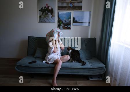 Müde Frau ruht sich auf der Couch aus mit Kissen über dem Kopf und einer Hauskatze neben ihr, überfordert und erschöpft zu Hause Stockfoto