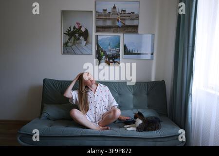 Glückliche Frau genießt das morgendliche Dehnen auf dem Sofa mit ihrer Katze, Entspannung, Wellness und bequemer Wohnstil. Stockfoto