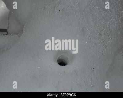 Nahaufnahme eines kreisförmigen Lochs, das durch eine dicke Schicht weißes oder hellgraues Material, wahrscheinlich Schnee, Eis oder verdichtetes Pulver, gebohrt oder geschmolzen wurde. Die Stockfoto