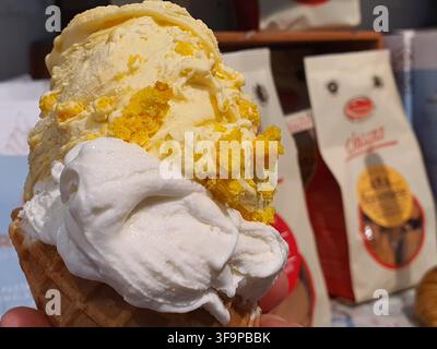 Nahaufnahme einer Hand, die ein Eis mit zwei Kugeln hält: eine cremige gelbe Kugel mit sichtbaren Stücken von gelbem Kuchen oder Keksstücken gemischt Stockfoto