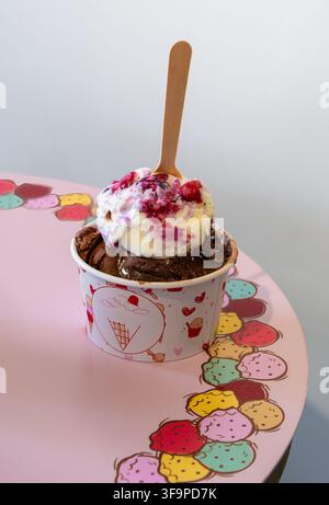 Löffel Schokoladen-, Vanille- und Beeren-Eis in einem Pappbecher mit einem Holzlöffel auf einem rosa Tisch. Süßes Dessert im Nahaufnahme mit weichem Licht und verspieltem Stockfoto