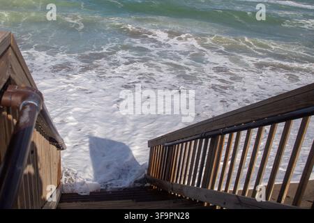 Eine Hochwinkelperspektive von der Spitze der hölzernen Strandzugangstreppe mit Blick nach unten auf Hochwasser-Ozeanwellen und weißen Schaum, der direkt auf das niedrige Ufer spült Stockfoto