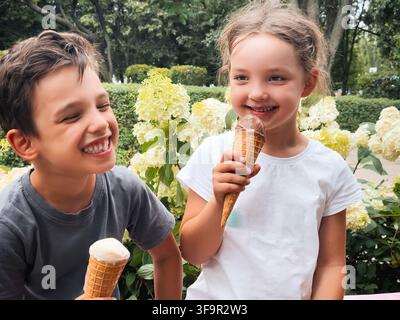 Zwei fröhliche Kinder, ein Junge und ein Mädchen, essen glücklich Eisbecher im Freien in einem üppigen grünen Park oder Garten mit blühenden hellen Blumen. Stockfoto