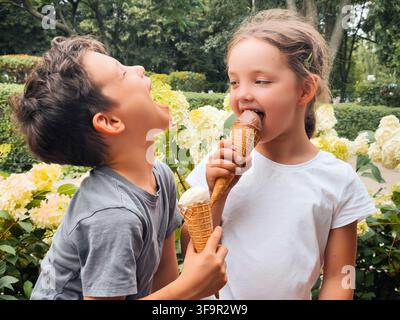 Ein fröhlicher Junge und ein Mädchen genießen Eiscremewaffeln in einer lebendigen Gartenszene mit blühenden weißen Blumen und halten einen entzückenden Moment des Sommers fest Stockfoto