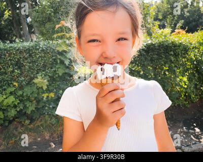 Fröhliches Mädchen lächelt, während es Vanille-Schokoladeneis im Freien isst. Glückliches Kind genießt ein süßes Dessert im hellen Gartenlicht. Kindheitsfreude co Stockfoto