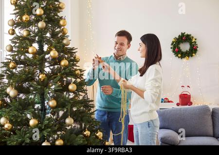 Fröhliches Paar dekoriert gemeinsam den Weihnachtsbaum mit Stern, Girlanden und anderen Ornamenten Stockfoto