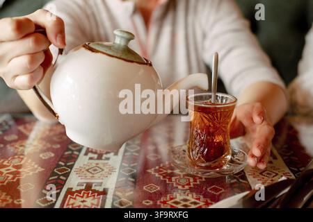 Hand hält eine Teekanne und gießt schwarzen Tee in eine türkische Tasse. Teetrinken in einem asiatischen Café Stockfoto