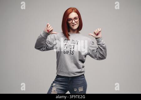 Ernte von modischen rothaarige Mädchen mit grauen Sweatshirt mit Beschreibung über für die ersten Geschenke. Stilvolle Frau posiert im Studio zeigen mit den Fingern Stockfoto