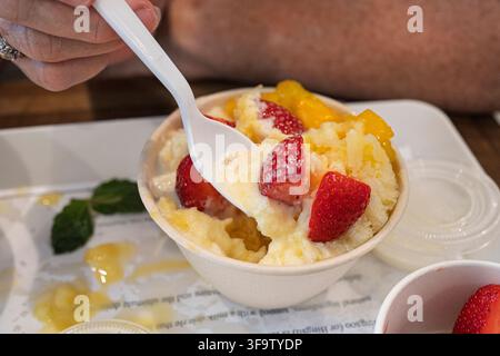 Hände greifen nach Schüsseln mit cremigem Mango-Geschabteis, das mit frischen Erdbeeren gekrönt ist und in Pappbechern auf einem weißen Tablett serviert wird. Diese zwanglose Essensszene fängt ein Stockfoto