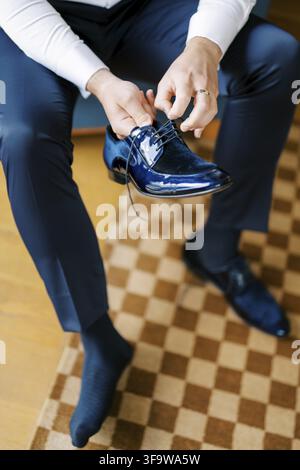 Ein Mann mit dunkelblauen Hosen und einem weißen Hemd bindet die Schnürsenkel von glänzenden blauen formellen Schuhen, während er auf einem karierten Teppich sitzt, mit einem Fuß in Stockfoto