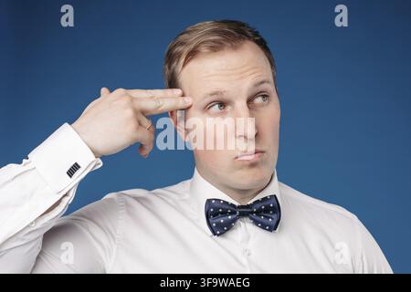 Vorderansicht des lustigen Mannes mit weißem Hemd und Bow Krawatte, die beiseite schauen und auf blauem isoliertem Hintergrund Pistolenzeichen zeigen. Junge männliche Gestiking und Stockfoto