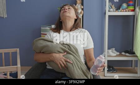 Frau, die ein eingewickeltes Baby und eine Flasche hält, Kopf nach hinten geneigt in einem Gebäude-Kinderzimmer; Müdigkeit Fürsorge Trost. Stockfoto