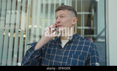 Mann, der das Smartphone mit der rechten Hand am Ohr hält, kariertes Hemd trägt, am Fenster des Bürogebäudes mit vertikalen Jalousien steht, während er telefoniert; Th Stockfoto