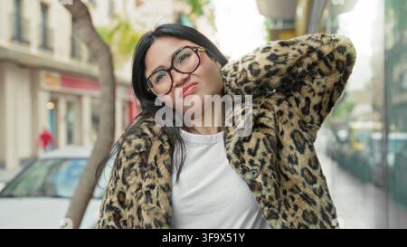 Frau mit Brille, die einen Leopardenprint-Mantel trägt, posiert mit der Hand im Nacken auf einer städtischen Straße neben einem geparkten Auto und einem Schaufenster, lächelt und neigt den Kopf; Stockfoto
