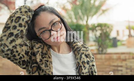 Frau mit Schildpatt-Brille in Leopardenmantel berührt Haare und lächelt auf einer Straße neben einer Ziegelmauer; zufriedener Charme. Stockfoto