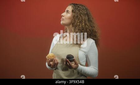 Junge Frau mit lockigen Haaren, die eine Schürze trägt und zwei Muffins hält, während sie sie in einem Studio mit rotem Hintergrund vergleicht; die Entscheidung über die Wahl ist durchdacht. Stockfoto