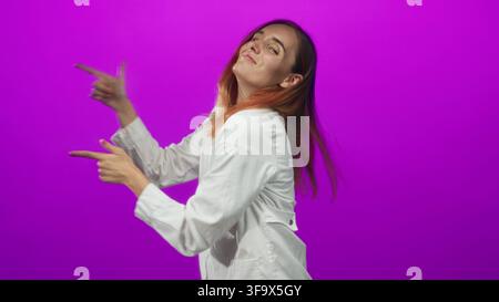 Frau Ärztin in weißem Mantel zeigt Finger wie Fingerpistolen im Studio, zeigt Hände und Zeigefinger zeigende Geste; spielerisches Selbstvertrauen. Stockfoto