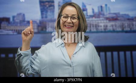 Reife hispanische Frau zeigt den Finger nach oben, lächelnd, zeigt Zeigefinger, trägt Brille und goldenen Anhänger im Studio; Vertrauensverbindung. Stockfoto