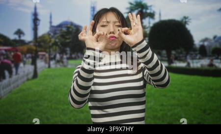 Junge Frau in gestreiftem Pullover macht mit beiden Händen ein OK-Zeichen und spitzt die Lippen, während sie auf grünem Rasen vor einem historischen Gebäude steht; verspielt Stockfoto