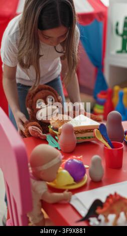 Mädchen, das ein ausgestopftes Affen- und Spielzeug-Sandwich auf einem roten Spieltisch zwischen Puppen und Plastikgeschirr im Gebäude platziert; spielerische Fantasie. Stockfoto