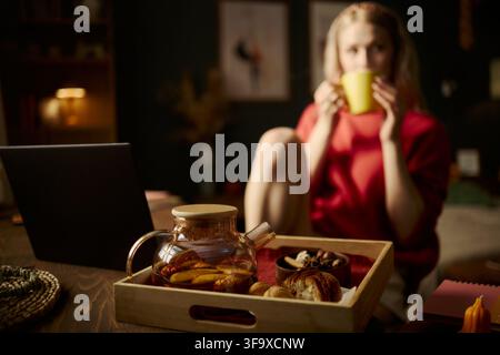 Teekanne auf Holztablett mit junger kaukasischer Frau im Hintergrund, die Tee trinkt und an einem Laptop arbeitet. Bild ideal für die Förderung von Telearbeit, Selbstfürsorge und gemütlichen Lebensstilkonzepten Stockfoto