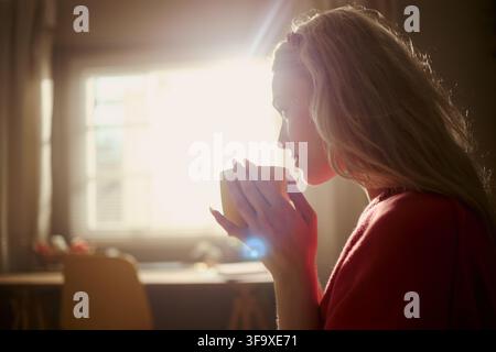 Junge kaukasische Frau hält eine Tasse und trinkt ein heißes Getränk zu Hause, Seitenansicht. Bild stellt Morgenroutine und Selbstfürsorge dar, geeignet für Kampagnen zum mentalen Wohlbefinden Stockfoto