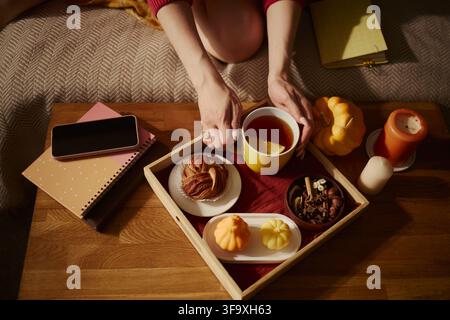 Kaukasische junge Frau, die eine gelbe Tasse mit heißem Tee auf einem Holzteller mit Herbstsnacks hält. Bild, das ein gemütliches Lebensstilkonzept für saisonale Marketingkampagnen darstellt, Hochwinkelansicht Stockfoto