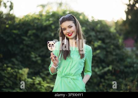 Porträt der Schönen lächelnde Mädchen im grünen Kleid, Spaß haben, in der Kamera candy auf Stick. Hübsche und stilvolle Frau mit roten Lippen, lange Ohrringe Stockfoto