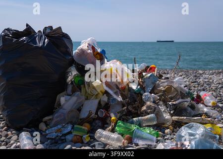 Großer Haufen von Plastikflaschen, Tüten und verschiedenem Müll neben einem vollen schwarzen Müllsack auf einem Kiesstrand, der marine und Umweltverschmutzung veranschaulicht Stockfoto