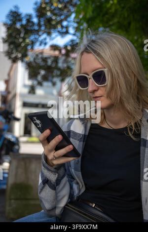 Junge Frau, die eine Sonnenbrille und ein kariertes Hemd trägt und ihr Smartphone im Freien benutzt. Sie repräsentiert digitale Konnektivität und moderne li Stockfoto