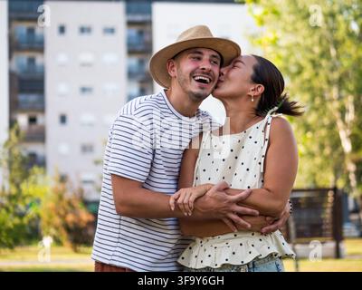 Fröhlicher Mann und Frau teilen eine herzliche Umarmung und einen Kuss, umgeben von Grün in einem Stadtpark. Stockfoto