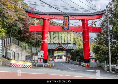 Ein großes rotes Torii mit der Inschrift „大天狗山“ markiert den Eingang zum Hakone Ōtengu-san-Schrein auf der alten Hakone-Straße zwischen Yumoto und Moto-Hakone, Präfektur Kanagawa, Japan. Steinerne Löwenhund-Wächter (komainu) stehen am Fuß der Säulen, während goldene Engelsfiguren auf dem Querbalken schweben. Der 1980 gegründete Schrein ist bekannt für mizuko kuyō-Gedenkrituale für verstorbene Kinder und verbindet Shinto mit westlicher Bildsprache. Stockfoto