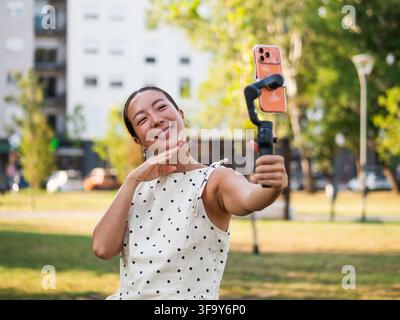 Lächelnde Content-Schöpferin filmt sich selbst mit einem Smartphone-Stabilisator an einem sonnigen Tag im Park. Stockfoto
