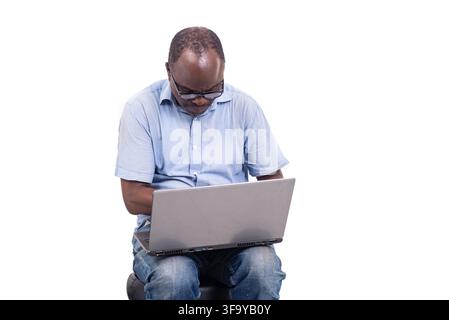 reifer Mann mit Brille, der auf einem Ottoman über weißem Hintergrund sitzt und an einem Laptop arbeitet. Stockfoto