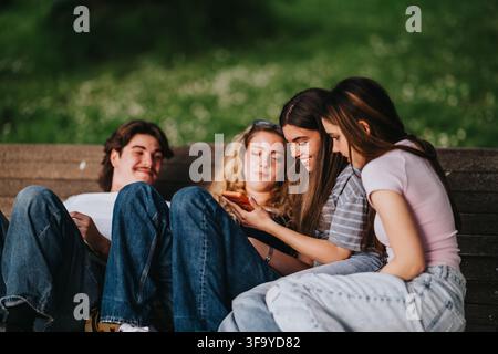 Vier Freunde sitzen auf einer Parkbank, lachen und schauen gemeinsam auf ein Smartphone Stockfoto