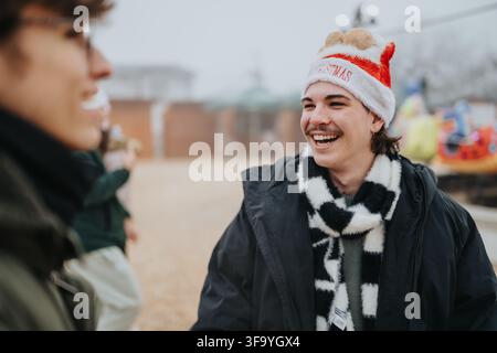 Junger Mann mit einem Weihnachtsmannhut lacht mit Freunden im Freien auf einem Wintervergnügungsgebiet Stockfoto