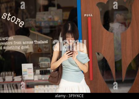 BANGKOK, THAILAND - 12. AUGUST 2026: Touristin nutzt ihr Smartphone, während sie vor einem Geschäft in Yaowarat steht, Vorderansicht. Stockfoto