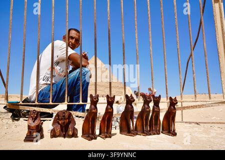 Ägyptischer Souvenirladenbesitzer sitzt hinter einem Metallzaun mit hölzernen Bastet-Katzen und Sphinx-Statuen in der Nähe der Großen Pyramide von Gizeh Stockfoto