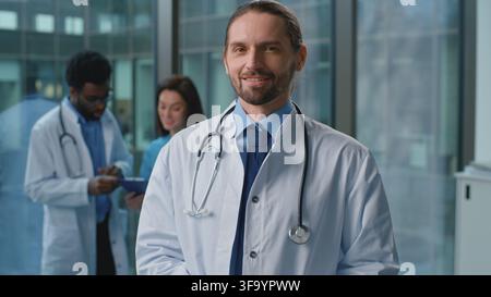 Positiv lächelnder kaukasischer Mann, Arzt, arbeitet in der Klinik und schaut in den Korridor in der Nähe von Kollegen, Ärzten, in die Kamera. Optimistischer männlicher Spezialist, Therapeut in Stockfoto