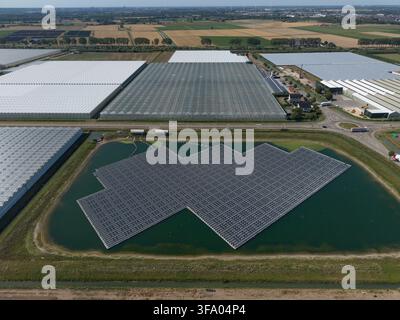 Luftaufnahme einer großen schwimmenden Solarzellenanlage auf einem Teich, gelegen neben weitläufigen Glashäusern und landwirtschaftlichen Feldern in den Niederlanden. Stockfoto