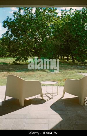 Blick aus einem Schlafzimmer auf eine kleine Terrasse mit einem Tisch und Stühlen, mit Blick auf Walnussbäume, die eine ruhige ländliche Szene mit einem heimischen Charme schaffen. Stockfoto