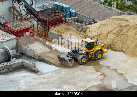 Luftaufnahme eines gelben Radladers, der in einer Zementfabrik in Polen mit Sand-, Kieshaufen und industriellen Fördergeräten arbeitet. Stockfoto