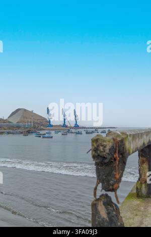 Blick auf Puerto Chancay mit industriellen Kränen und kleinen Fischerbooten im Hafen, gesehen von einem verwitterten Betonpier in Chancay, Lima Region, Peru Stockfoto