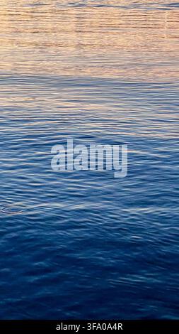 Ruhige blaue Wasseroberfläche mit sanften Wellen und warmen goldenen Reflexionen, die einen friedlichen abstrakten Naturhintergrund erzeugen. Stockfoto