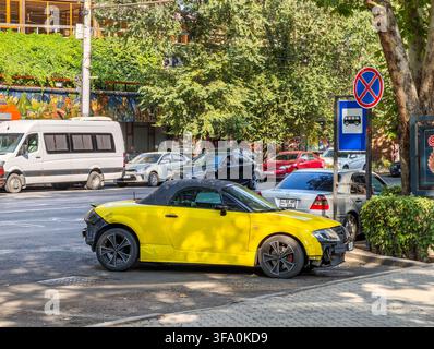 Gelbes Cabriolet, das auf einer Kopfsteinpflasterstraße geparkt ist. Bäume und geparkte Autos umgeben die Szene. Blaues Parkschild in der Nähe des Fahrzeugs sichtbar. Sonnenlichtfilter Stockfoto