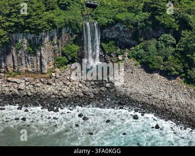 Luftbild des Jeongbang-Wasserfalls, der einen felsigen Abhang hinunter in das Meer stürzt, umgeben von üppigen grünen Bäumen und einem Kiesstrand in Südkorea. Stockfoto